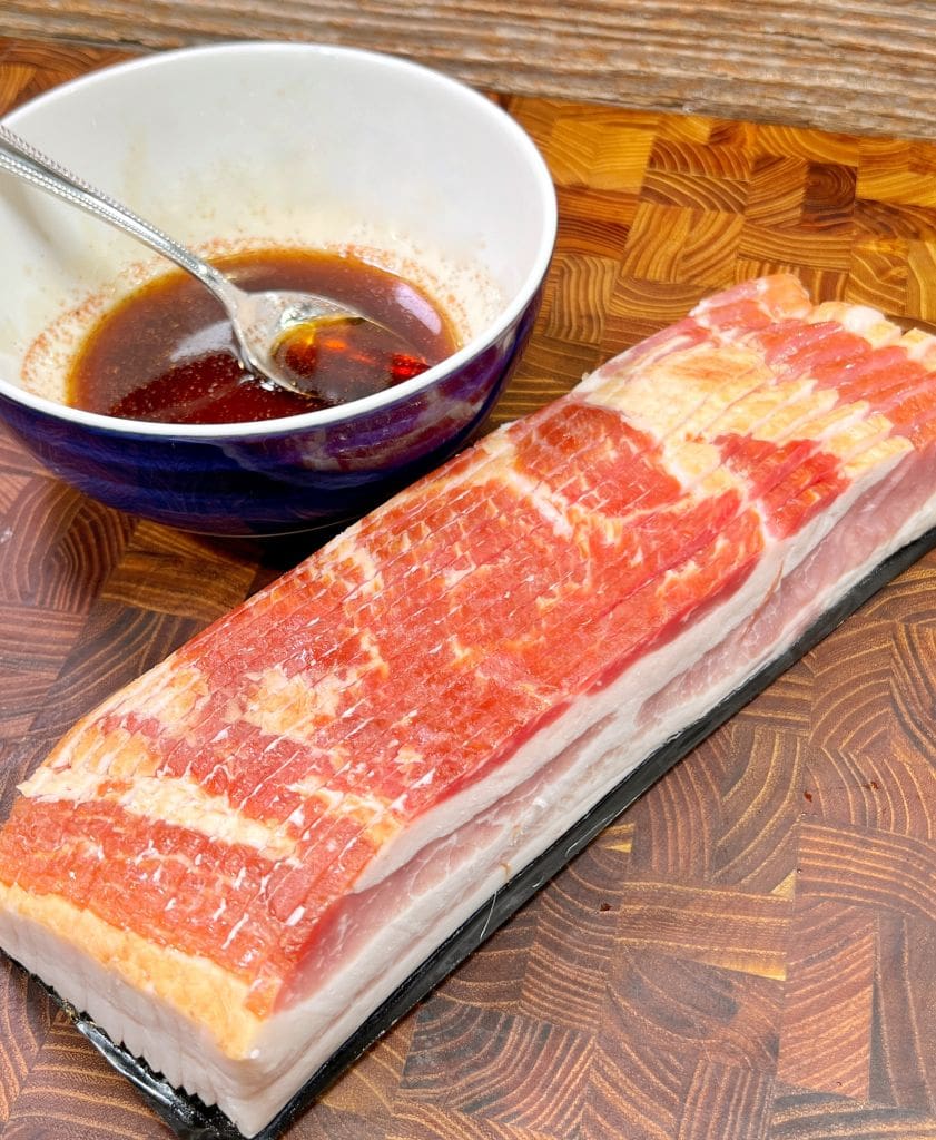 A slab of raw bacon on a wooden cutting board next to a bowl containing a red marinade with a spoon in it.