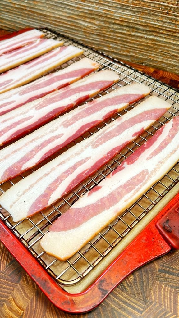 Strips of raw bacon are evenly spaced on a metal cooling rack placed on a red baking sheet. The setup is ready for oven cooking, resting on a wood-patterned surface.