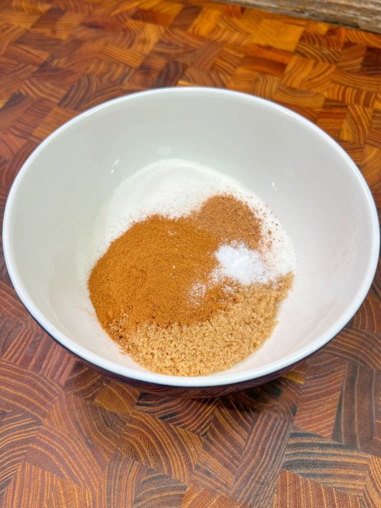 A white bowl containing ingredients for baking on a wooden surface. Inside are white sugar, brown sugar, cinnamon, and a small amount of salt.