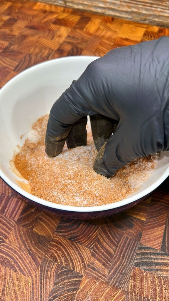 A gloved hand mixes a blend of spices in a white bowl set on a wooden surface. The spices include a mix of brown and white powders.