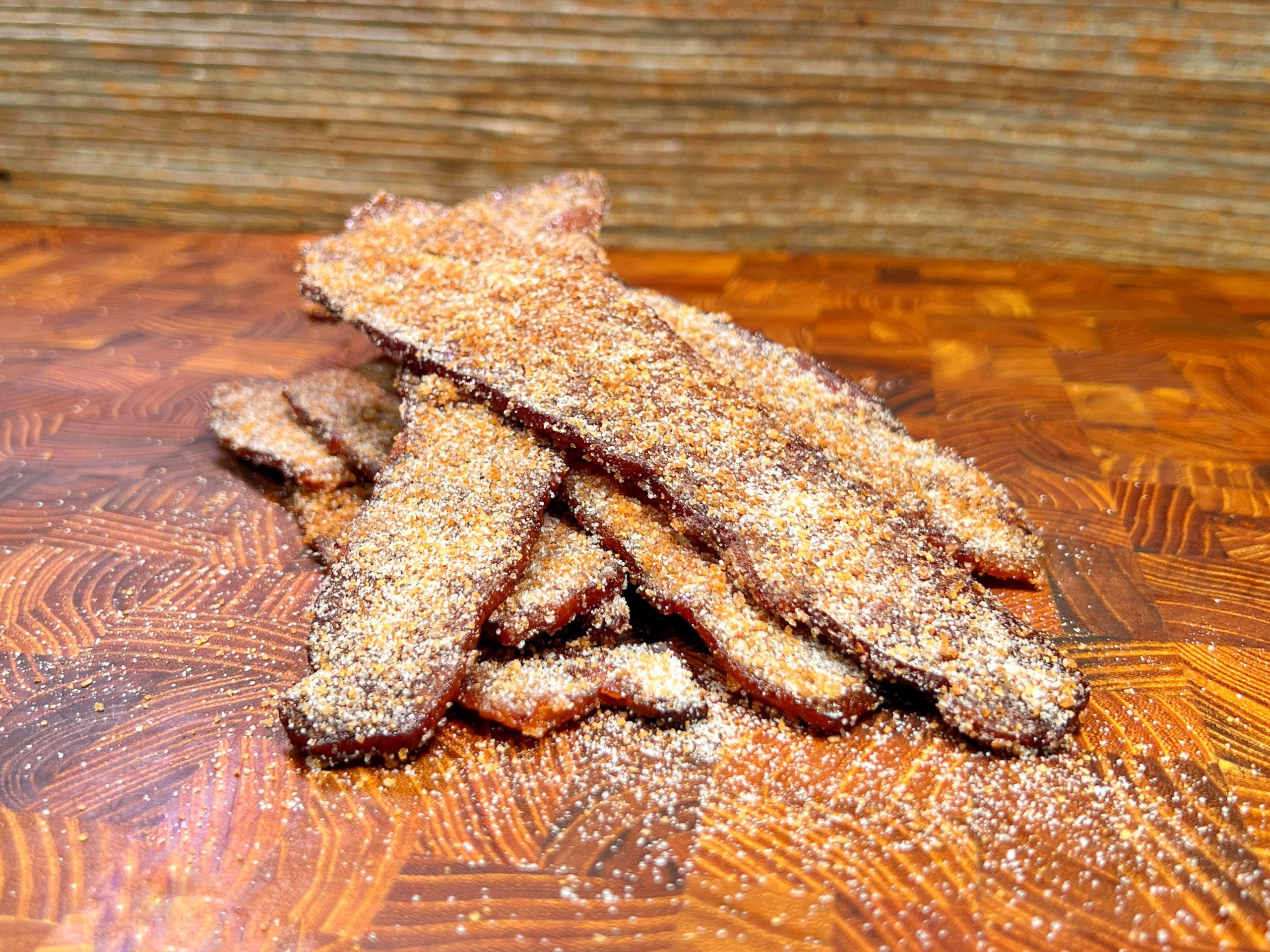 A pile of crispy bacon strips coated with a grainy seasoning sits on a wooden cutting board with a textured pattern. The background is made of horizontal wooden planks.