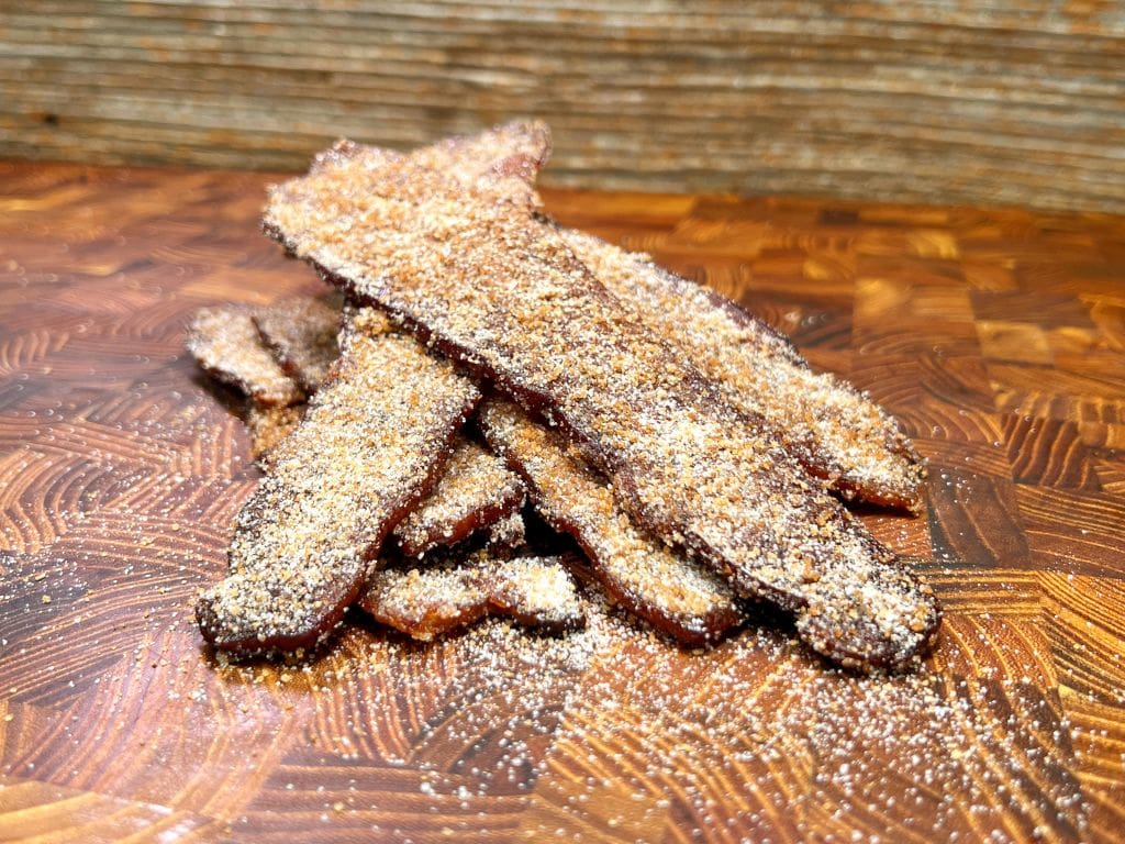 A stack of crispy, churro candied bacon strips is piled on a textured wooden surface with a rustic background. The bacon is heavily coated with a sugar mixture, emphasizing its caramelized edges and crunchy appearance.