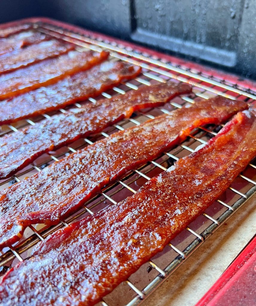 Close-up of several strips of candied bacon placed evenly on a wire rack. The glossy, crispy texture suggests it's been perfectly cooked. The wire rack rests over a red tray or baking sheet, giving the sweet-savory treat an appetizing display.