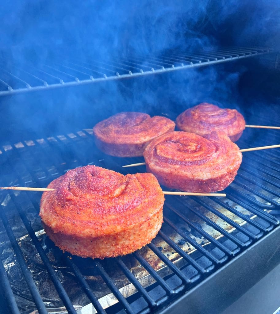 Four spiraled smoked pork belly skewers are cooking on a grill with smoke wafting around them. The meat has a rich reddish-brown color, likely seasoned with spices. The grill grates and the smoky atmosphere suggest outdoor cooking or barbecue.