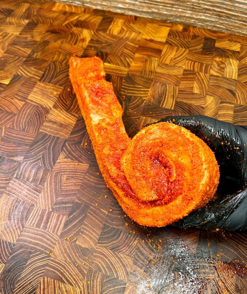 A raw pork belly strip, rolled and seasoned with red spices, is held by a gloved hand on a wooden cutting board with a chevron pattern.