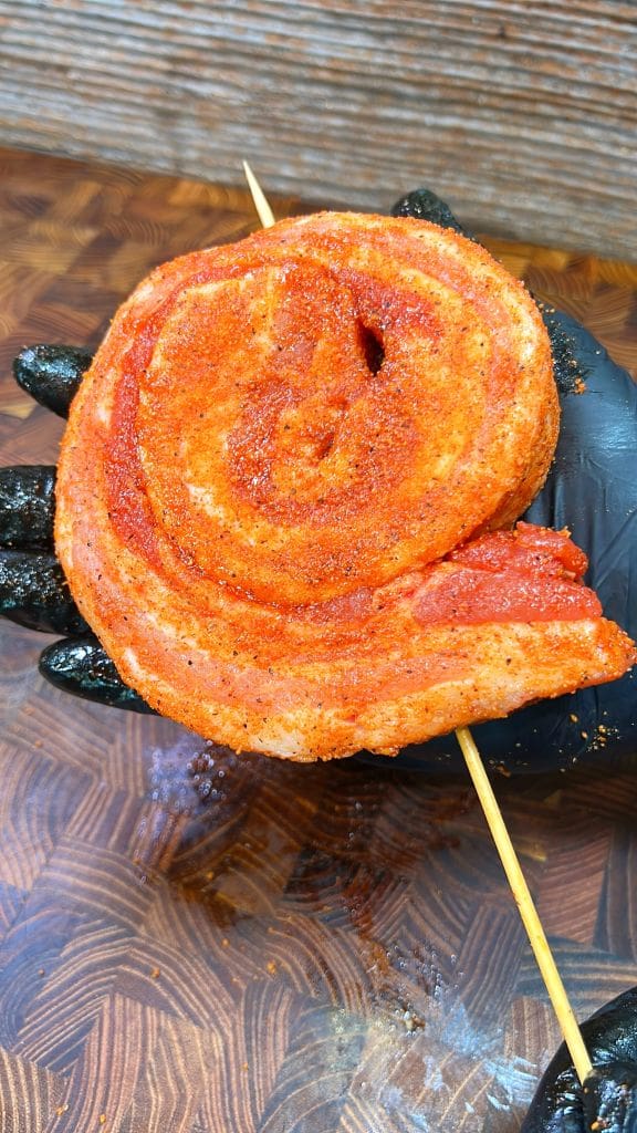 A gloved hand holds a rolled slice of seasoned smoked meat on a skewer, with a dark wooden background. The meat appears to be well-coated with spices, giving it a reddish hue.