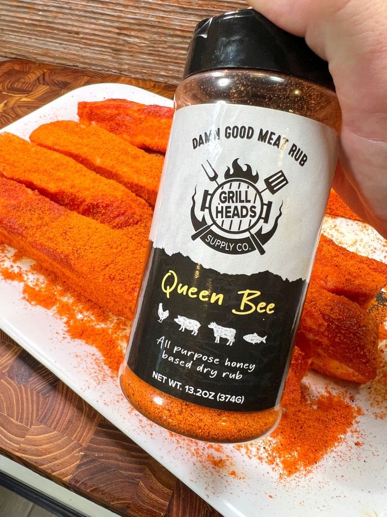 A hand holds a container of Queen Bee all-purpose honey-based dry rub by Grill Heads, with red seasoning visible inside. In the background, ribs are generously coated with the rub on a white plate.