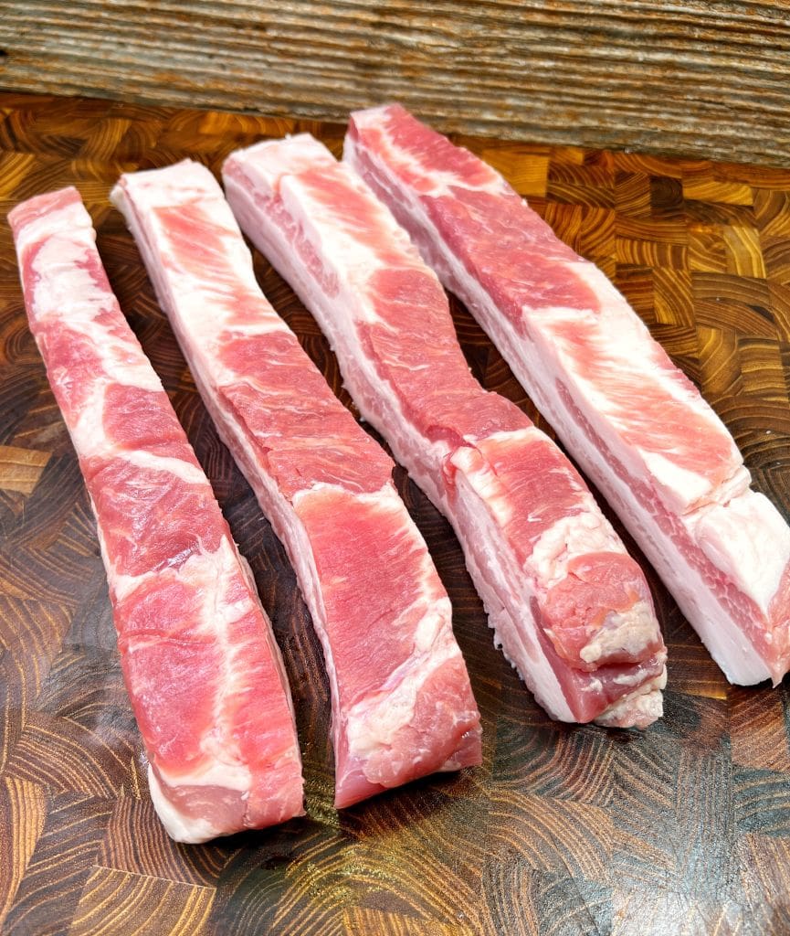 Four raw pork rib strips are placed on a wooden cutting board. The meat is pink with white marbling and cuts of fat, set against a textured wood backdrop.