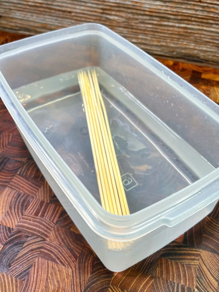 A plastic container filled with water and wooden skewers soaking inside. The container is placed on a wood-grain surface.