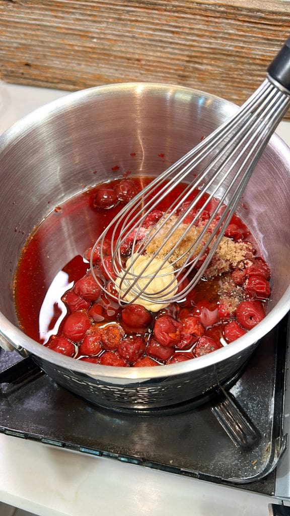 A metal pot on a stovetop contains cherries, brown sugar, and a pat of butter being mixed with a whisk. The ingredients are starting to melt and combine into a sauce.