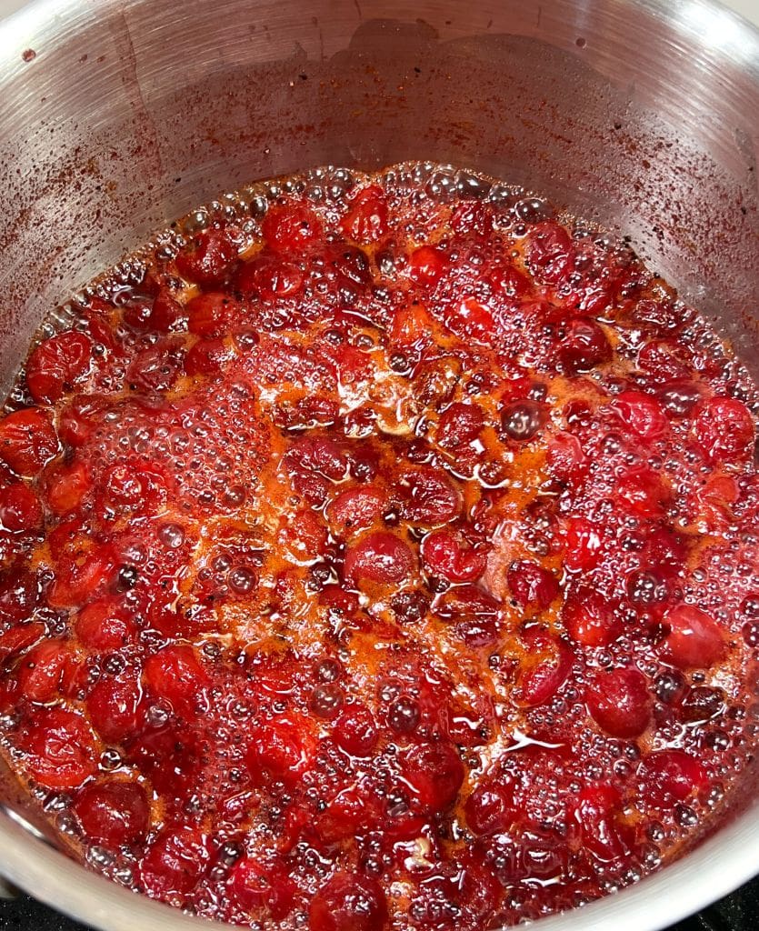 A pot filled with bubbling cranberry sauce being cooked. The cranberries are partially submerged in a thick, red liquid, creating a glossy and textured appearance. The mixture is slightly frothy around the edges.