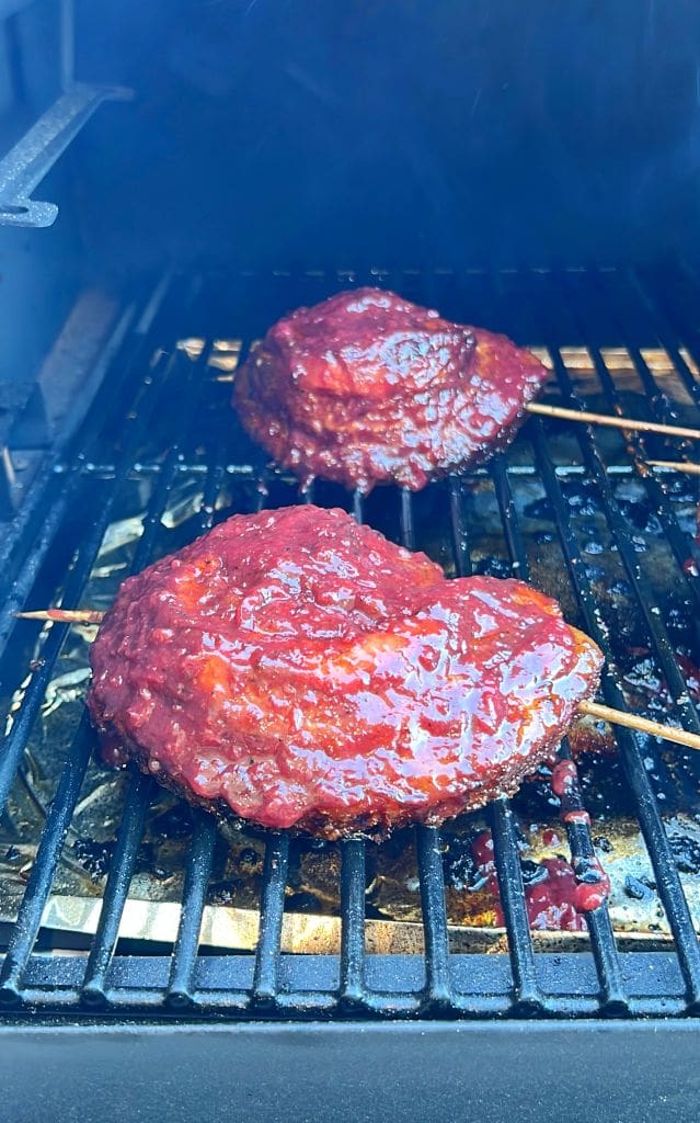 Two pieces of smoked pork belly coated in a rich red glaze are cooking on a grill inside a smoker. Skewers are visible beneath the meat, and smoke surrounds the scene.