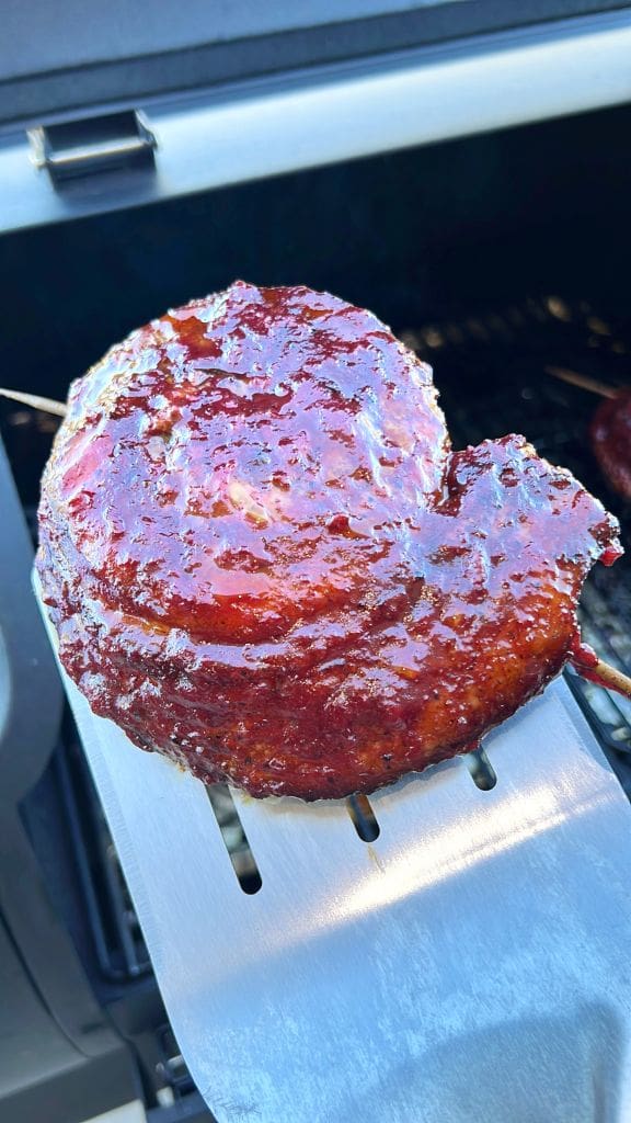 Close-up of a piece of barbecued meat with a shiny, caramelized glaze, resting on a metal spatula. The background shows an outdoor grill.