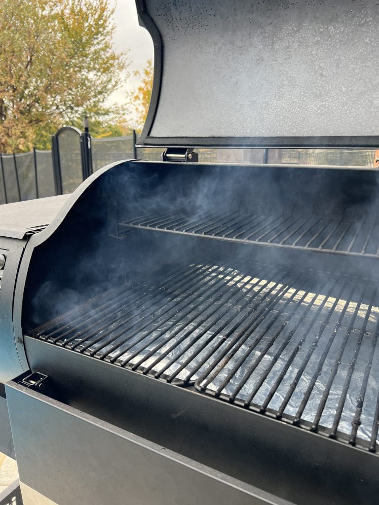 Smoker grill with open lid, emitting smoke. The clean grates eagerly await sizzling strips of bacon, set against a backdrop of a cloudy sky and trees.