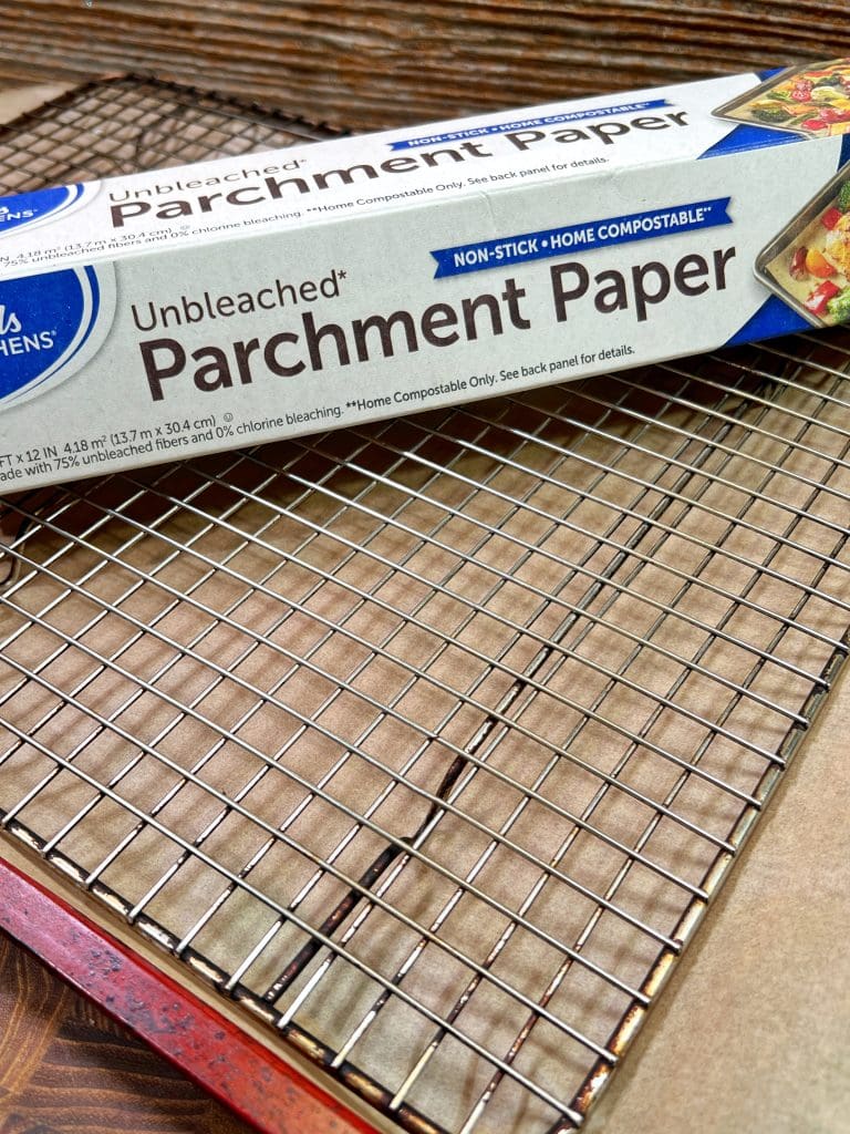 A box of unbleached parchment paper labeled Non-stick, Home Compostable sits on a wire cooling rack. The packaging displays a colorful dish with crisply cooked bacon, highlighting its use for non-stick cooking or baking needs.