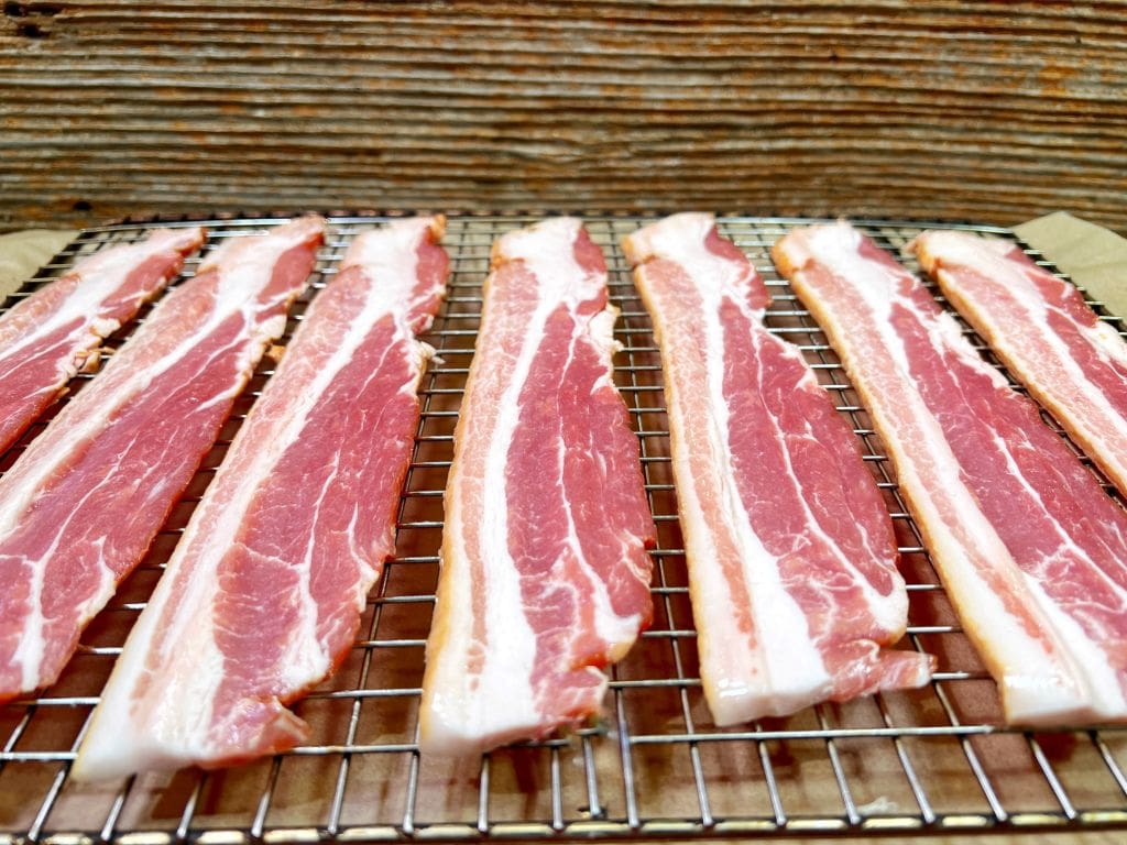 Eight slices of raw bacon are evenly spaced on a metal rack over a baking sheet. The background features wooden planks, adding a rustic touch to the scene.