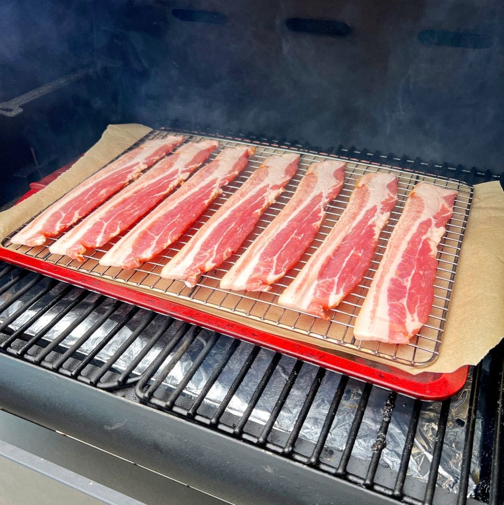 Uncooked bacon strips on a wire rack inside a smoker. Smoke surrounds the meat, and a red tray is placed underneath the rack to catch drippings. Brown parchment paper lines the tray.