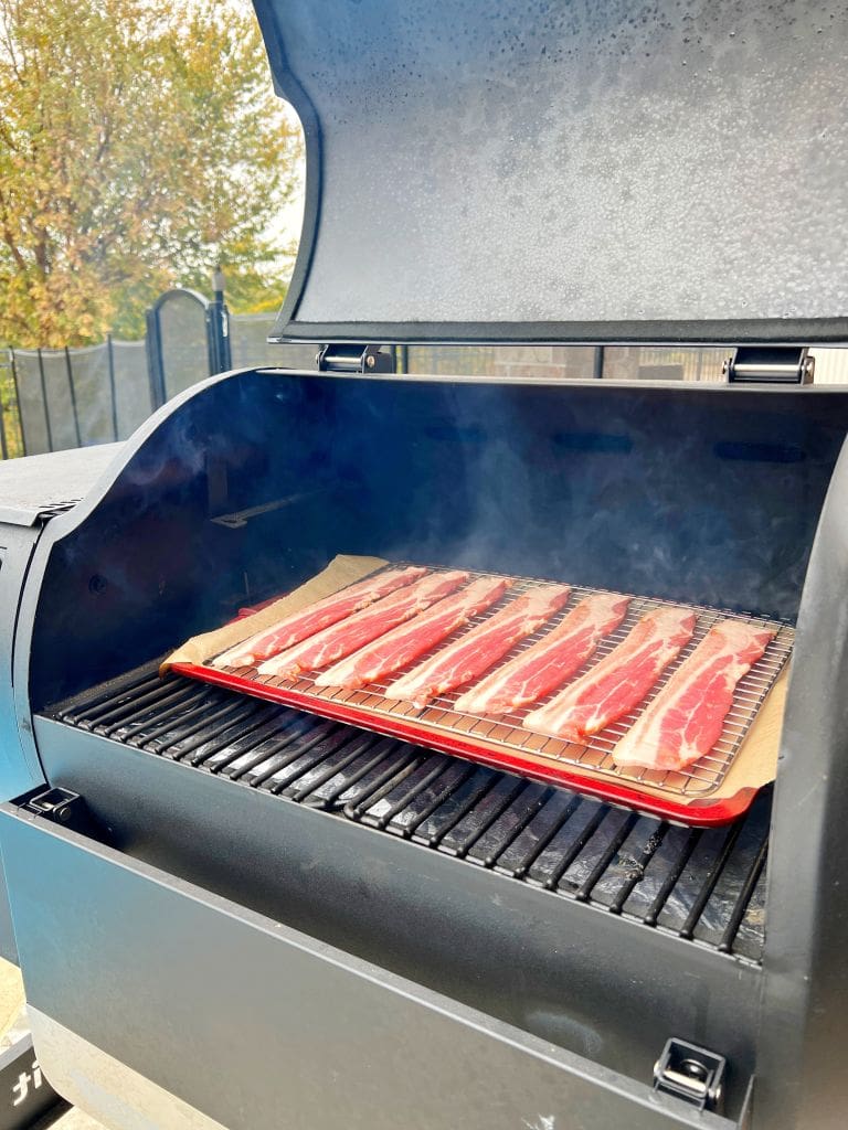 Outdoor grill with an open lid showing strips of bacon cooking on a tray inside. Smoke is visible, and a fence and tree are in the background.