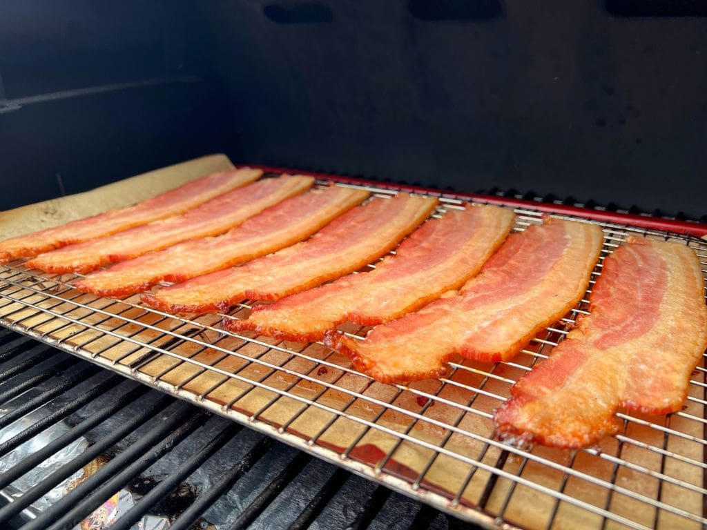 Slices of bacon cooking on a grill, placed neatly on a wire rack for even heat distribution. The bacon is sizzling and appears close to being perfectly crispy, set against a dark grill background.