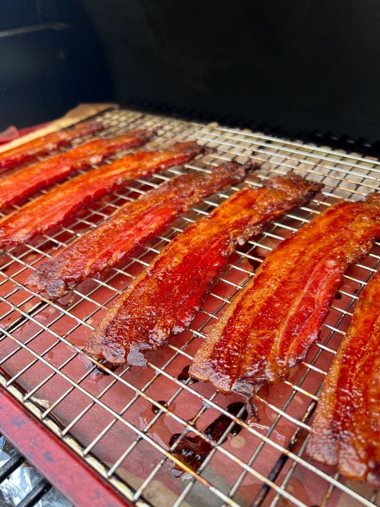 Close-up of several strips of crispy bacon sizzling on a grill rack. The bacon is a rich golden brown with visible fat and slight charring. The grill beneath the rack is dark, providing contrast to the vibrant color of the bacon.