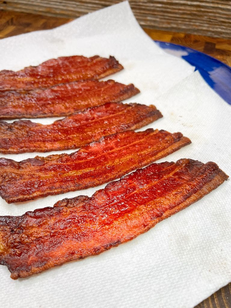 Four crispy strips of bacon resting on white paper towels, placed on a wooden surface. The bacon has a rich, red-brown color, indicating its well-cooked. A hint of a blue plate is visible on the side.