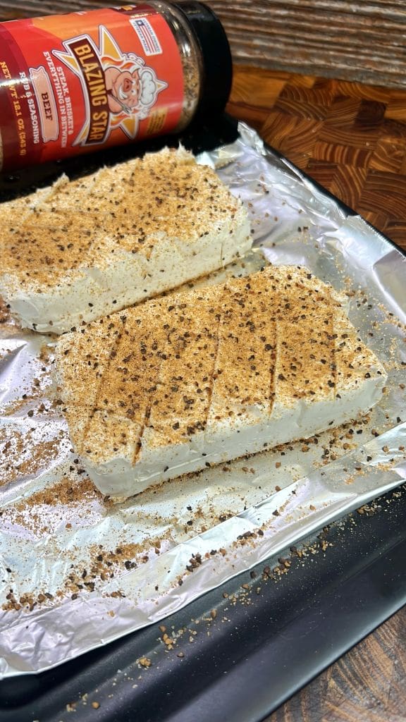 Two blocks of cream cheese on a foil-lined baking tray, topped with a seasoning mix. The seasoning bottle is in the background, labeled Blazing Star. The cream cheese blocks have cuts on the top surface.