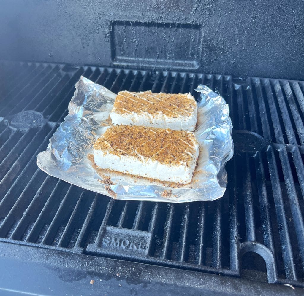 Two seasoned tofu blocks on a piece of aluminum foil placed on a grill, with grill marks visible on the tofu.