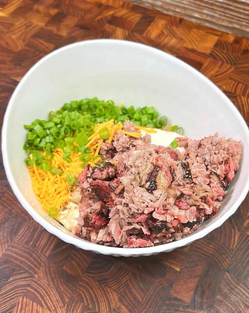 A white bowl containing a mix of shredded brisket, shredded cheddar cheese, chopped green onions, and mashed potatoes on a wooden surface.