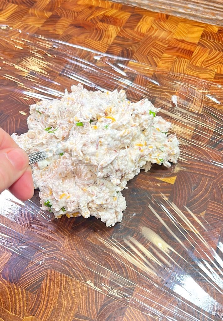 A person arranges a mixture of shredded chicken, cream cheese, cheddar, bacon, and herbs on clear plastic wrap set on a wooden surface for the cheeseball recipe. The mixture is about to be wrapped and formed into a ball.