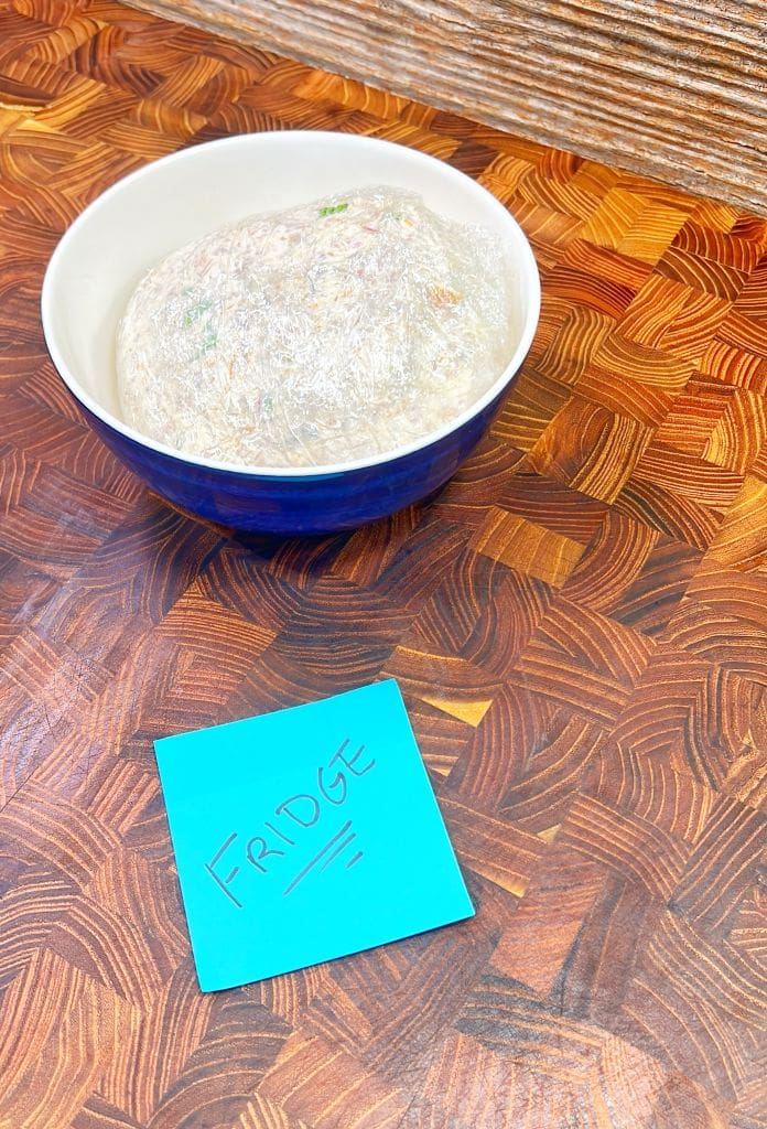 A blue bowl covered with plastic wrap sits on a wooden surface next to a blue sticky note with FRIDGE written on it.