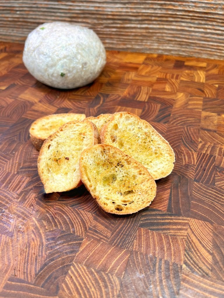 Slices of toasted baguette with herbs are arranged on a wooden cutting board. In the background, a round loaf wrapped in white plastic wrap is visible. The wooden board has a unique pattern.