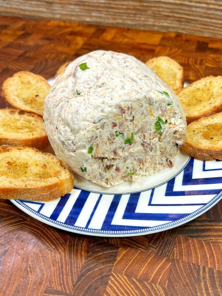 A round cheese ball with chopped herbs and nuts is placed on a blue and white plate. It is surrounded by slices of toasted baguette on a wooden surface.