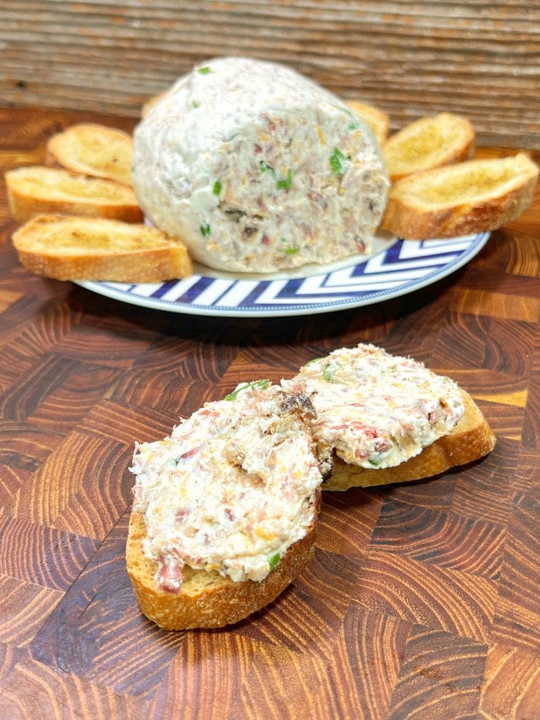 A dish of creamy cheese dip with herbs and bits of bacon, surrounded by toasted bread slices. Two slices with spread are placed in the foreground on a wooden surface. The dip is on a blue and white patterned plate.