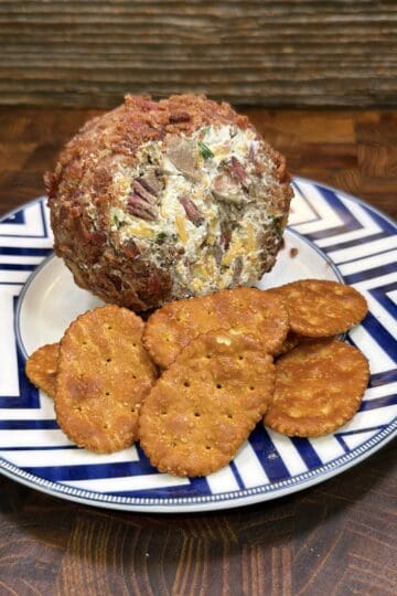 A round cheeseball, made from a classic cheeseball recipe and coated with pecans and herbs, is served on a plate with several seasoned crackers, set on a wooden surface.