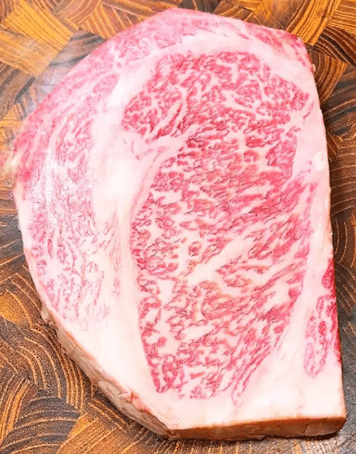 A close-up of a raw steak with rich marbling patterns, showcasing a blend of red meat and white fat. The wooden cutting board beneath contrasts with the intricate marbling.