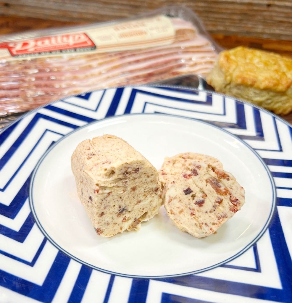 A round block of bacon-flavored butter is cut in half on a white plate with a blue geometric border. In the background, theres packaged bacon and a biscuit on a wooden surface.