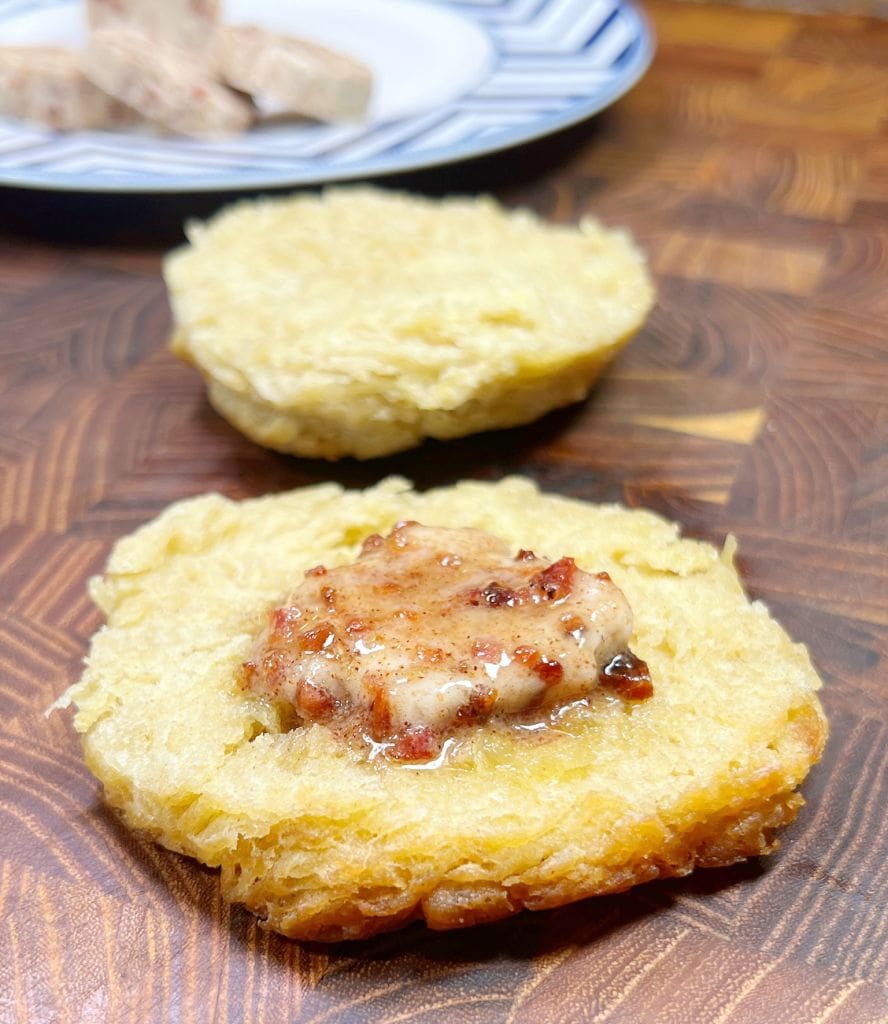 A toasted biscuit topped with a spreadable patty sits on a wooden surface. In the background, a plate with additional patties is partially visible.