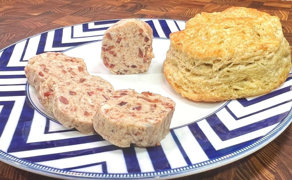 A plate with several slices of compound butter with visible bacon bits next to a golden-brown biscuit, placed on a blue and white patterned plate on a wooden table.