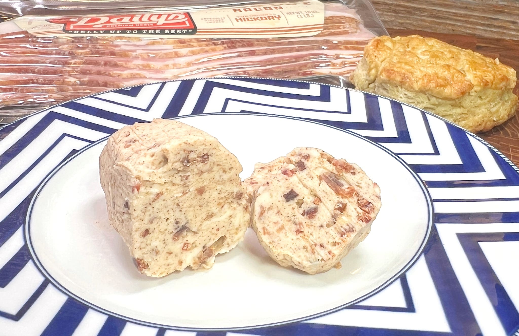 A plate with two slices of bacon-infused butter sits on a zigzag-patterned dish. Behind it, a package of hickory bacon and a biscuit are placed on a wooden table.
