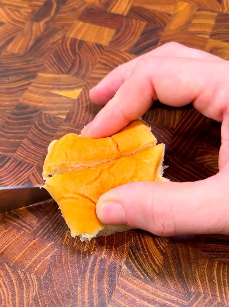 A hand slicing a small bun on a wooden cutting board with a knife. The background features a patterned surface.