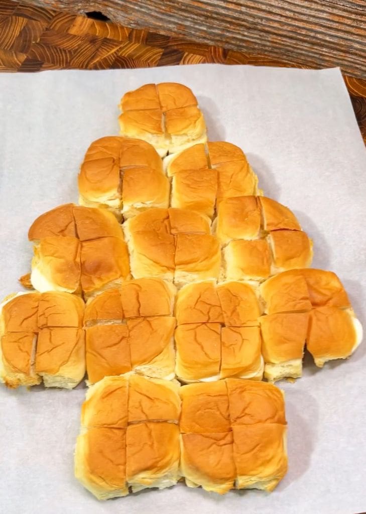 A group of dinner rolls arranged on parchment paper in the shape of a Christmas tree, with the top roll forming the trees peak. The rolls have a golden-brown crust.