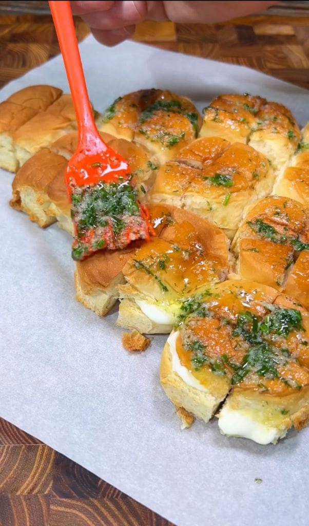 A person is using a red spatula to spread garlic herb butter on top of freshly baked bread rolls, some of which are topped with melted cheese. The rolls are placed on white parchment paper.