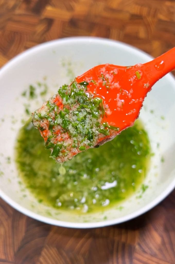 A red spoon holds a mix of chopped cilantro, garlic, and oil over a white bowl filled with the same green, herby mixture. The background features a wooden surface.
