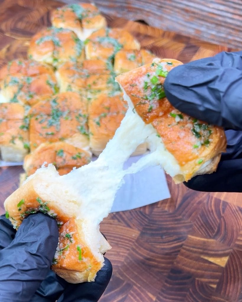 Gloved hands pulling apart soft, golden-brown garlic bread with stretchy, melted cheese, topped with herbs, on a patterned wooden surface. A tray of similar bread pieces is in the background.