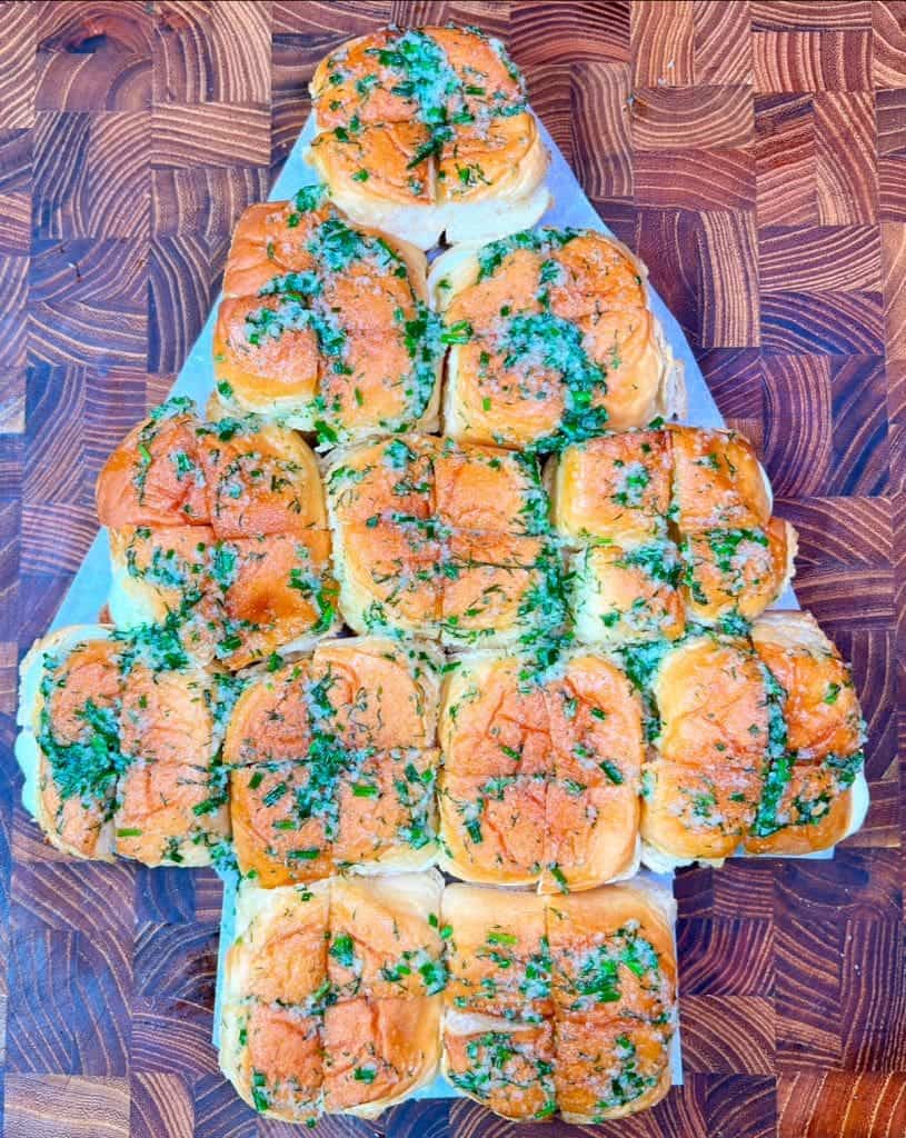 A Christmas recipe tree-shaped arrangement of golden-brown garlic bread rolls, garnished with chopped green herbs, sits on a wooden cutting board with a geometric pattern.