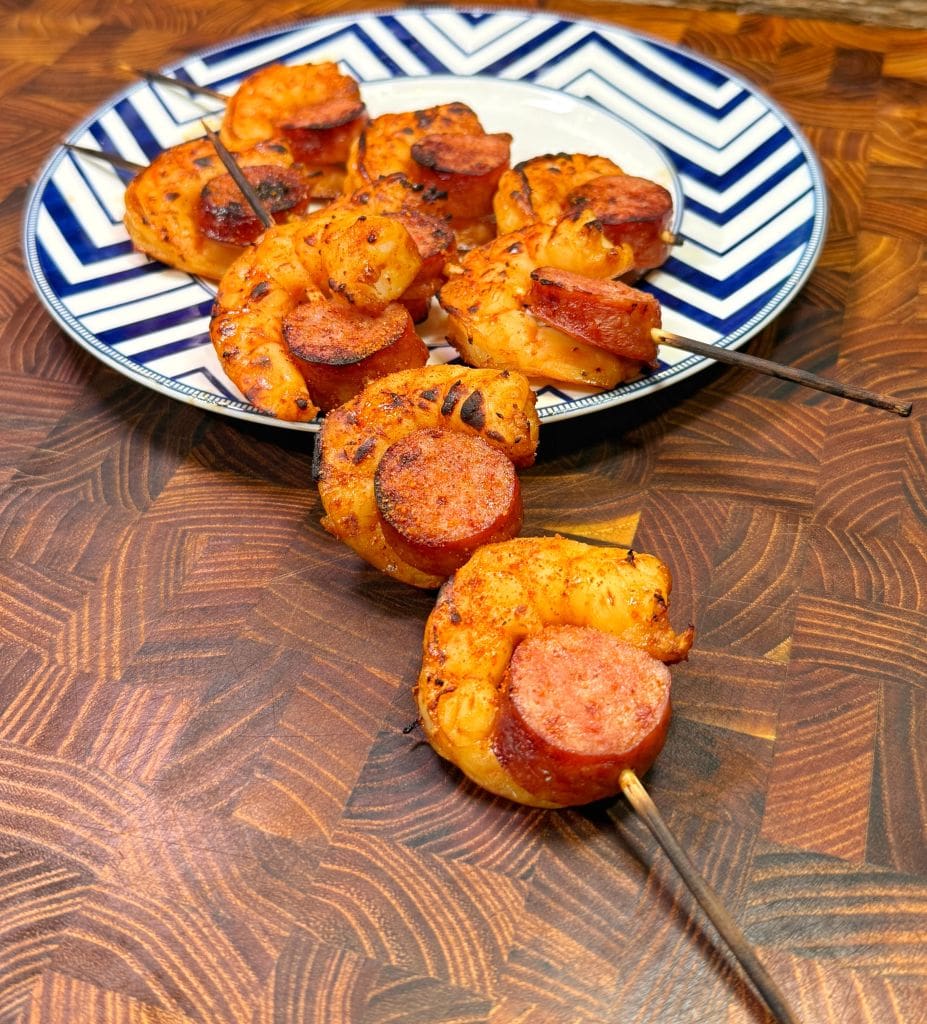 Grilled shrimp and sausage skewers on a decorative blue and white plate, placed on a wooden surface. The skewers are seasoned and cooked, showing a golden-brown texture.