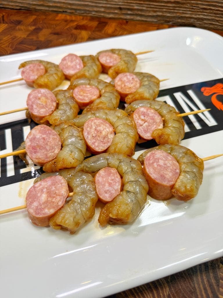 Raw shrimp and sausage slices are skewered alternately on sticks, arranged on a rectangular white plate with a decorative pattern.