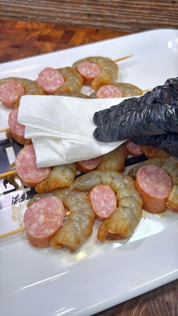 A gloved hand holding a paper towel is patting dry skewers with raw shrimp and sausage slices on a white plate. The background features a wooden surface.