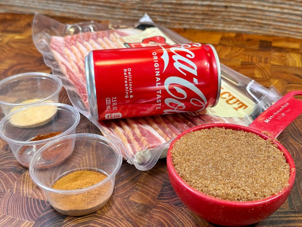 A can of Coca-Cola, a package of bacon, three small containers of spices, and a measuring cup filled with brown sugar are arranged on a wooden surface.