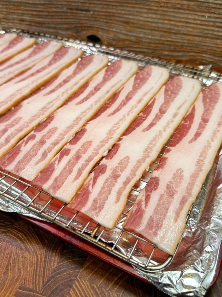 Several strips of raw bacon are arranged in a single layer on a wire rack covered with foil. The rack is placed on a wooden surface, ready for cooking. The bacon has visible marbling of fat and lean meat.